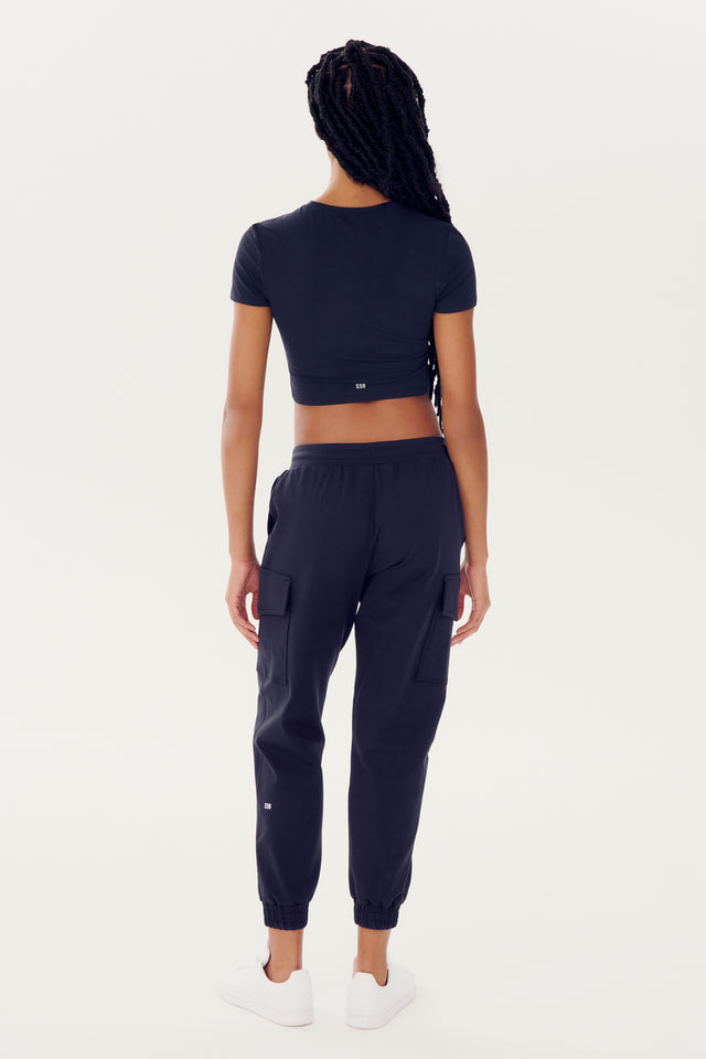 Woman standing facing away from the camera, wearing a quick dry SPLITS59 Airweight S/S Crop in indigo, navy joggers, and white sneakers.