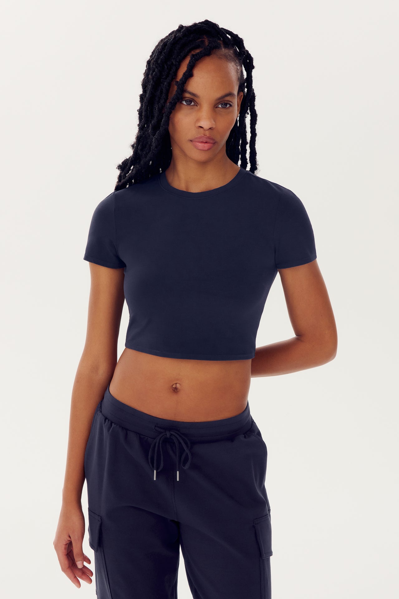 A woman in a black SPLITS59 Airweight S/S Crop - Indigo and sweatpants posing against a white background.