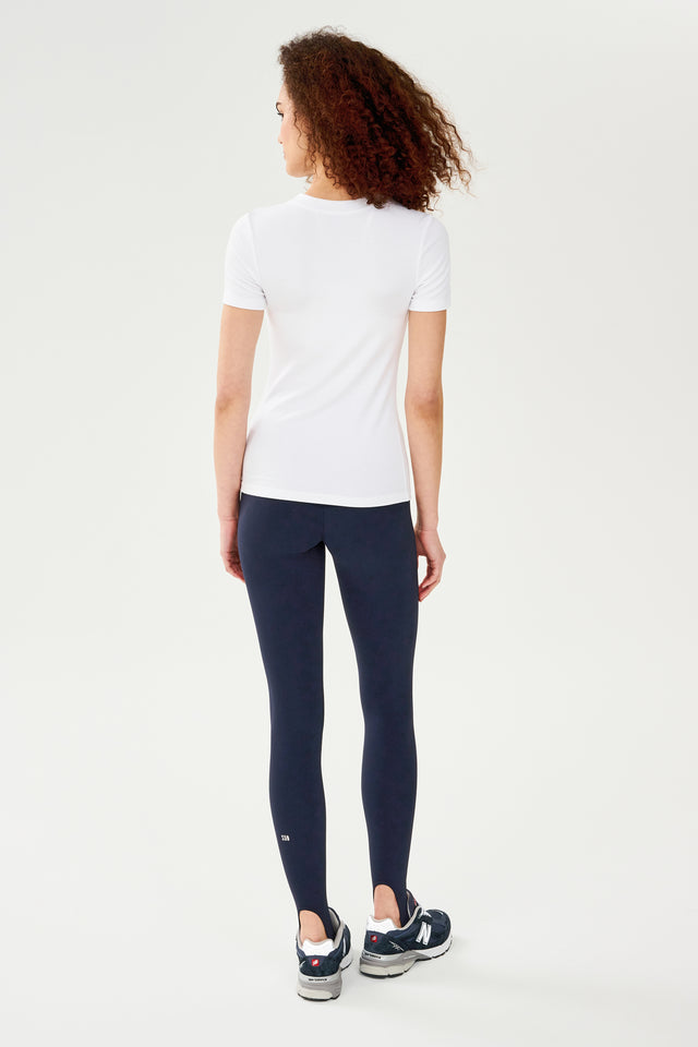 The back view of a woman wearing a SPLITS59 Louise Rib Short Sleeve - White t-shirt and navy leggings, ready for gym workouts.