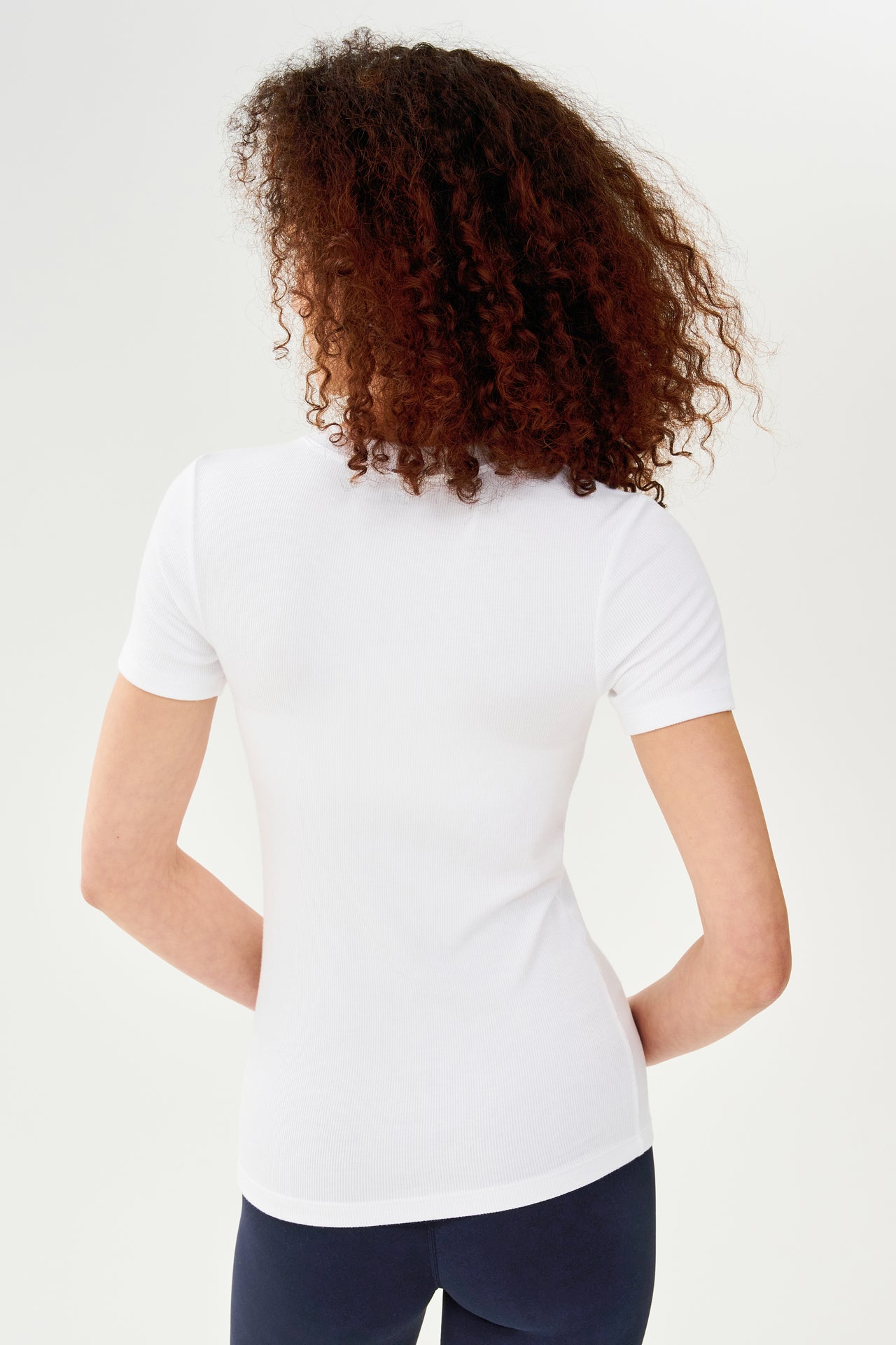 The back view of a woman wearing a SPLITS59 Louise Rib Short Sleeve - White t-shirt and leggings, ready for her yoga session.