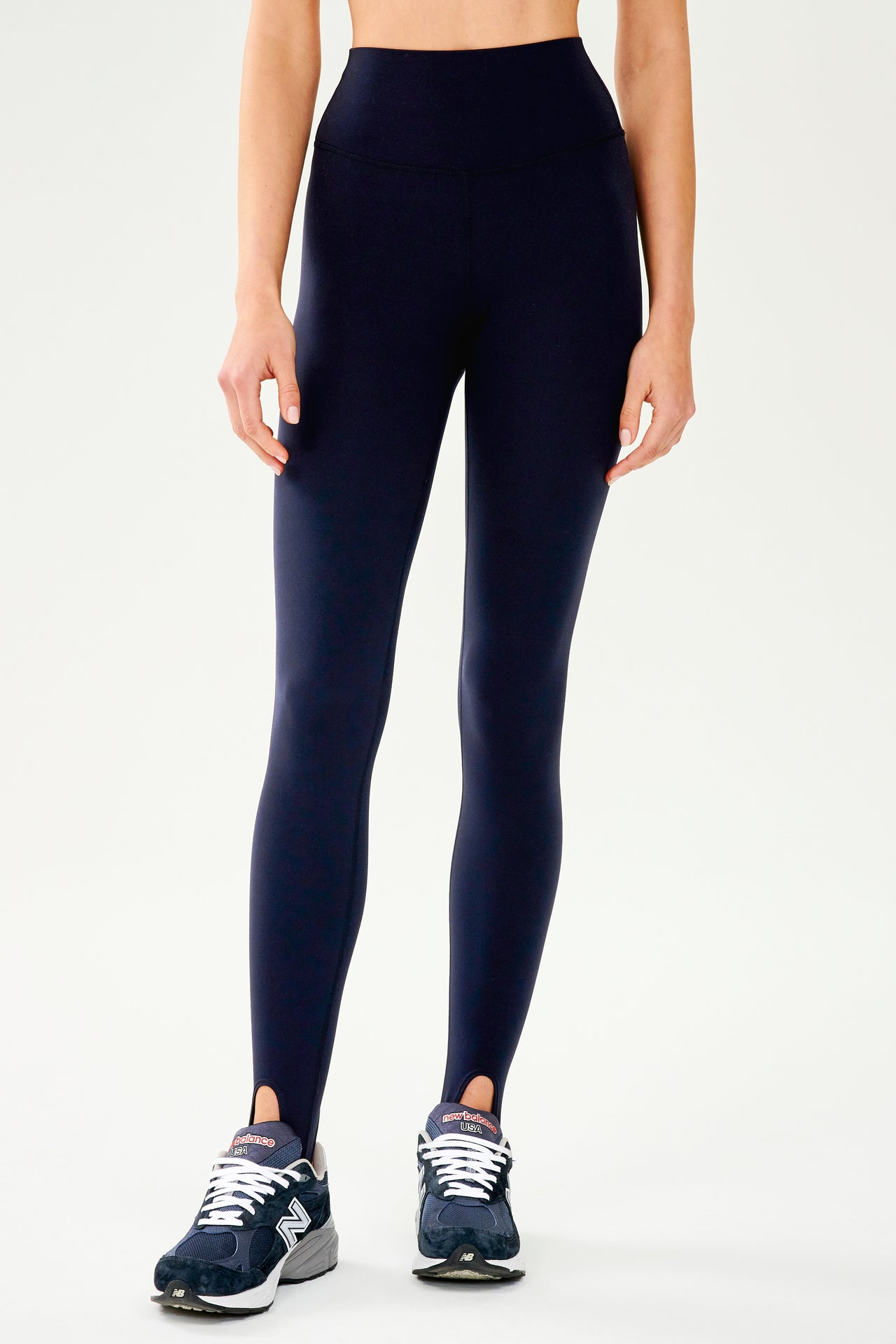 Front view of dark blue leggings with bottom strap that wraps around the foot paired with dark blue and white shoes