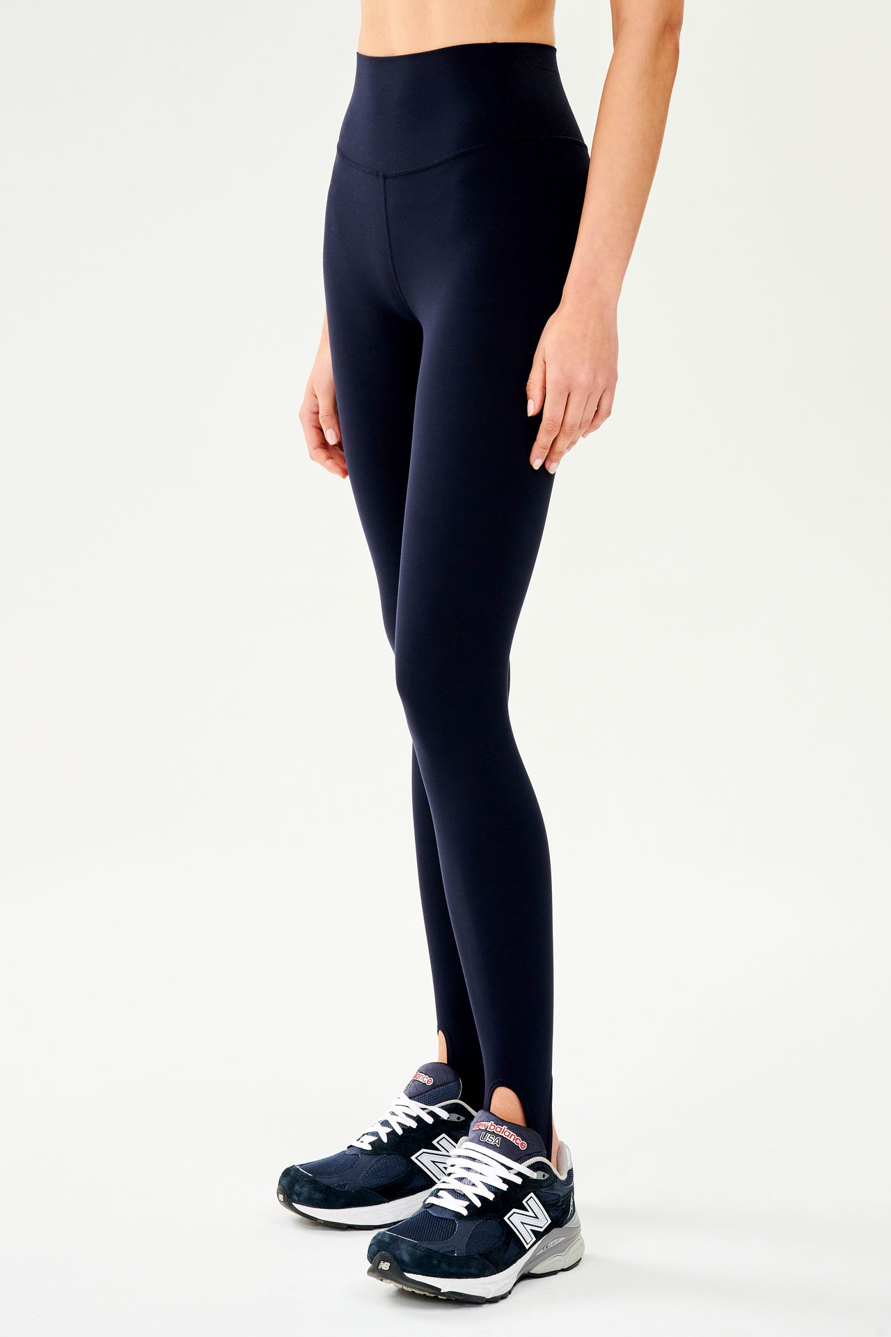 Front side view of dark blue leggings with bottom strap that wraps around the foot paired with dark blue and white shoes