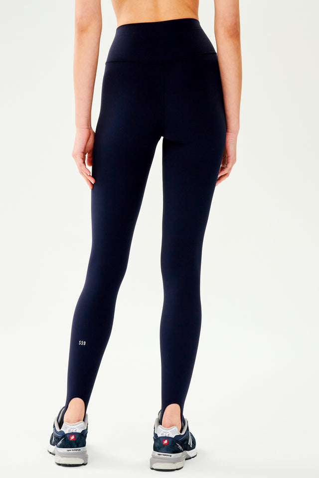 Back view of dark blue leggings with bottom strap that wraps around the foot paired with dark blue and white shoes