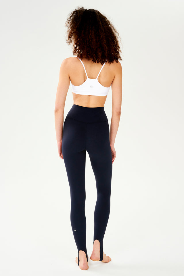 Full front side view of woman with dark curly hair wearing white bra with spaghetti racerback strap and dark blue leggings with bottom strap that wraps around the foot