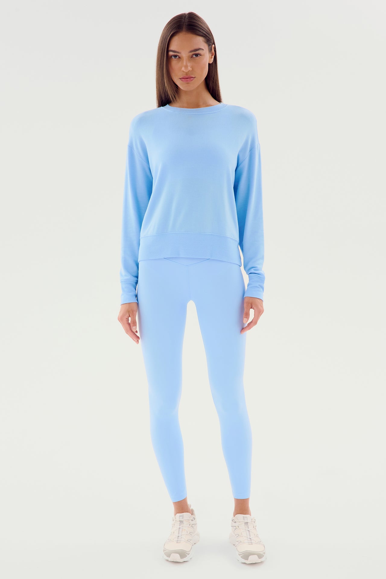 A woman stands facing forward, wearing the Sonja Fleece Sweatshirt in Skylight and matching light blue leggings made from ultra luxe fleece, paired with white sneakers against a white background.