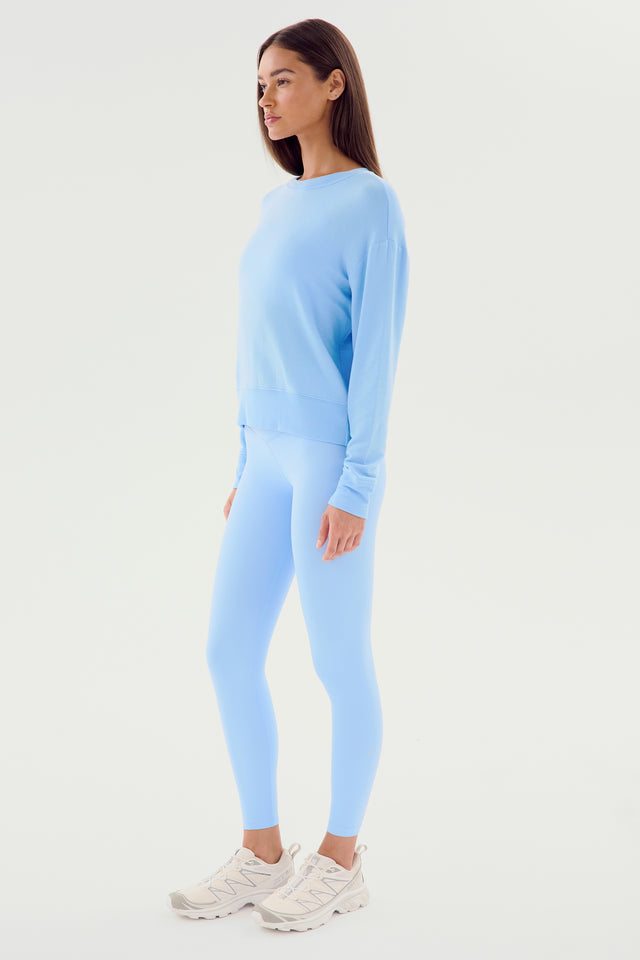 A person stands in profile, wearing a light blue Sonja Fleece Sweatshirt - Skylight by SPLITS59 with matching light blue leggings and white sneakers. The ultra-luxe fleece fabric of the outfit adds a touch of comfort and style. The background is plain and white, emphasizing the minimalist design MADE IN LOS ANGELES.