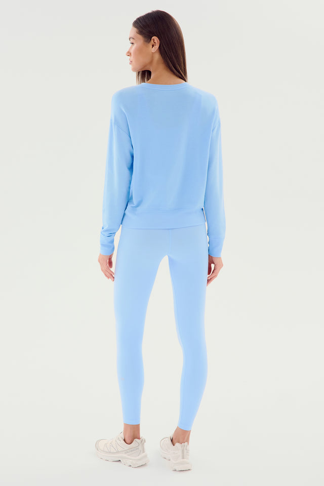 A woman is standing and facing away, showcasing the Sonja Fleece Sweatshirt in Skylight from SPLITS59 paired with matching blue leggings and white sneakers. The background is plain white. This chic outfit, MADE IN LOS ANGELES, epitomizes effortless style and comfort.