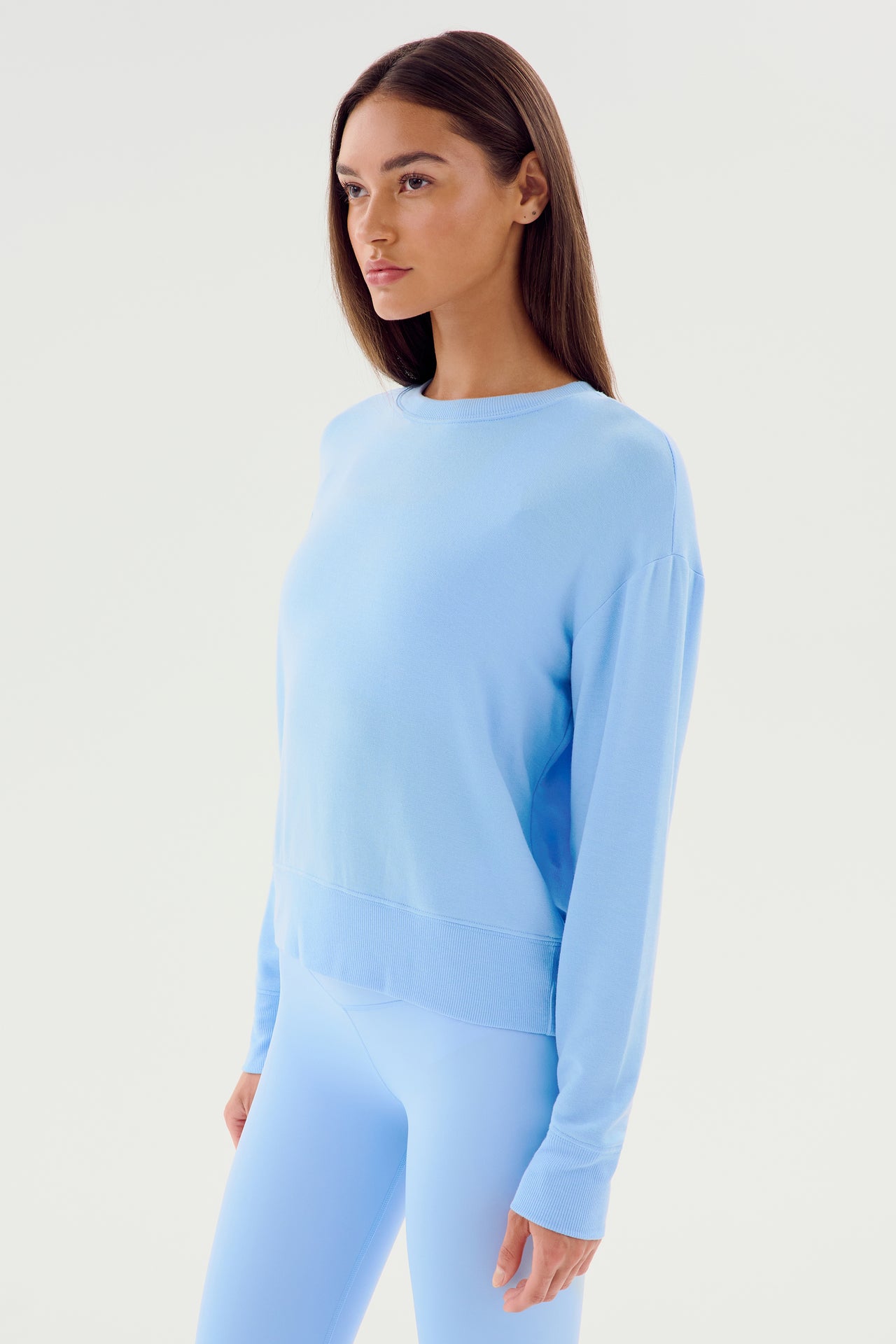 A person stands against a plain background wearing the ultra-flattering Sonja Fleece Sweatshirt - Skylight by SPLITS59 and matching light blue leggings. MADE IN LOS ANGELES, the outfit highlights a light blue hue. They are looking slightly to the side with a neutral expression.