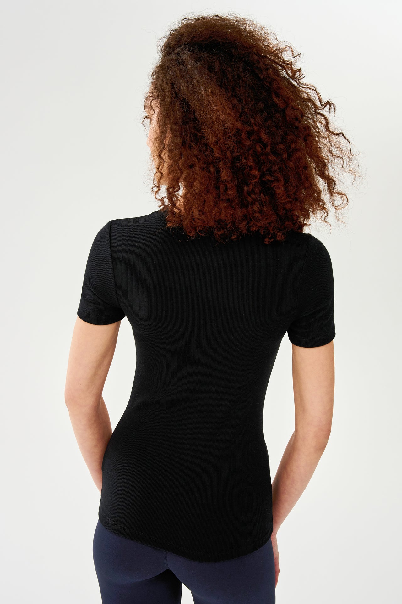 The back view of a woman wearing a SPLITS59 Louise Rib Short Sleeve - Black and leggings, ready for her yoga session.