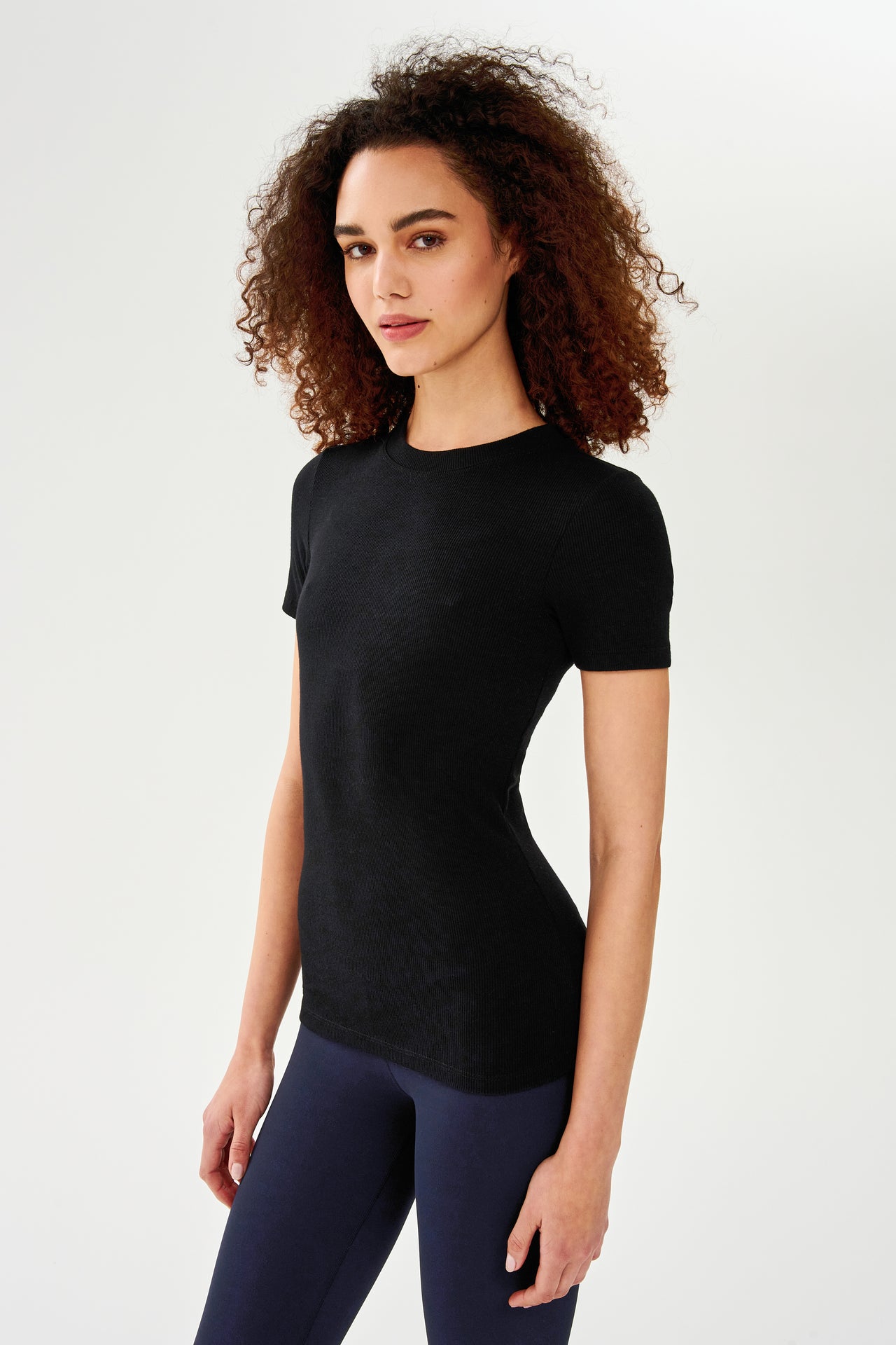 A woman wearing a SPLITS59 Louise Rib Short Sleeve - Black t-shirt and blue leggings, ready for her yoga session.