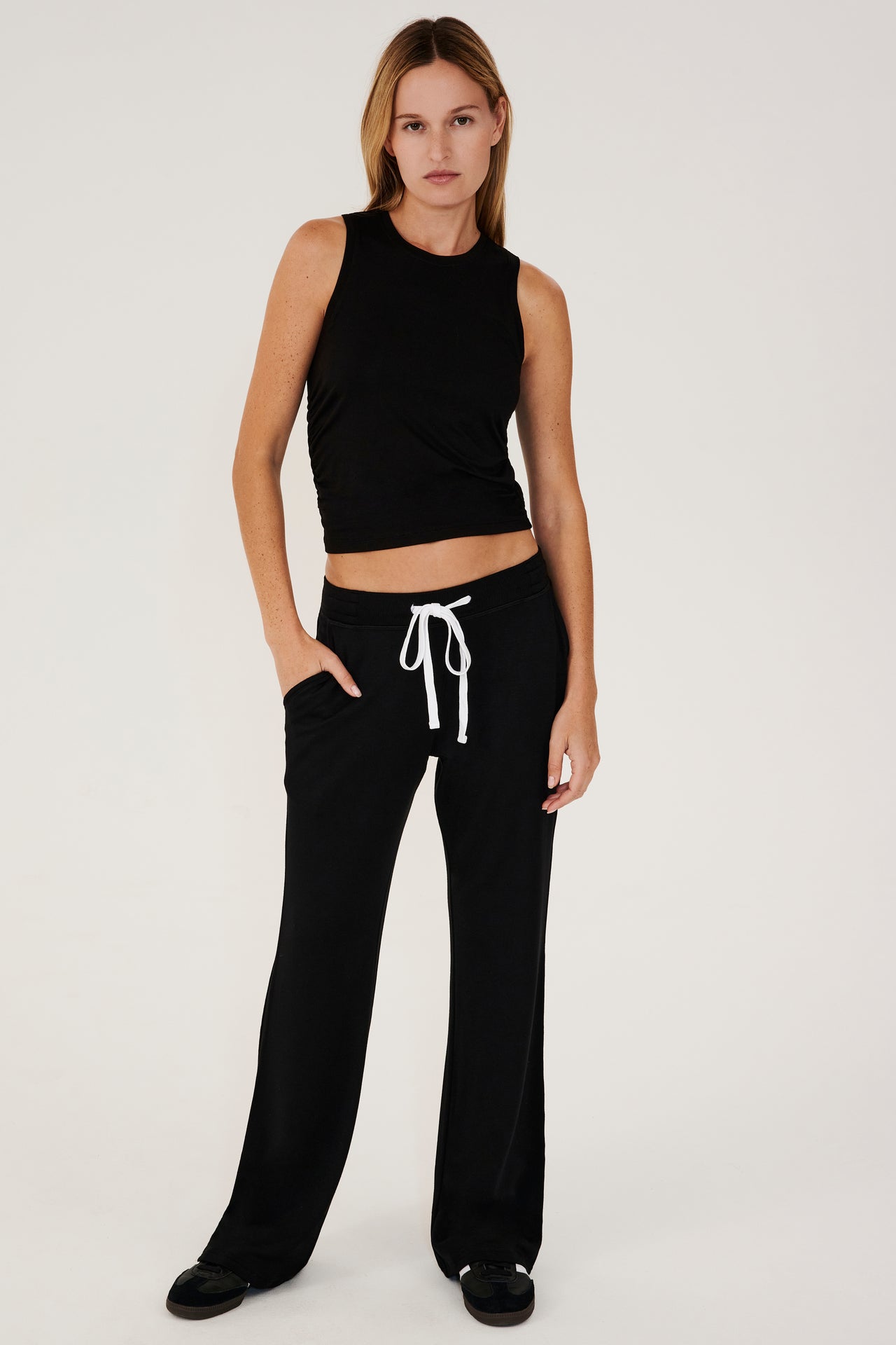 Full front view of woman with straight blonde hair wearing a black high rise wide leg relaxed fit sweatpant with side pockets and white drawstring. With black  high neckline racer tank top. Paired with black shoes with white stripes.