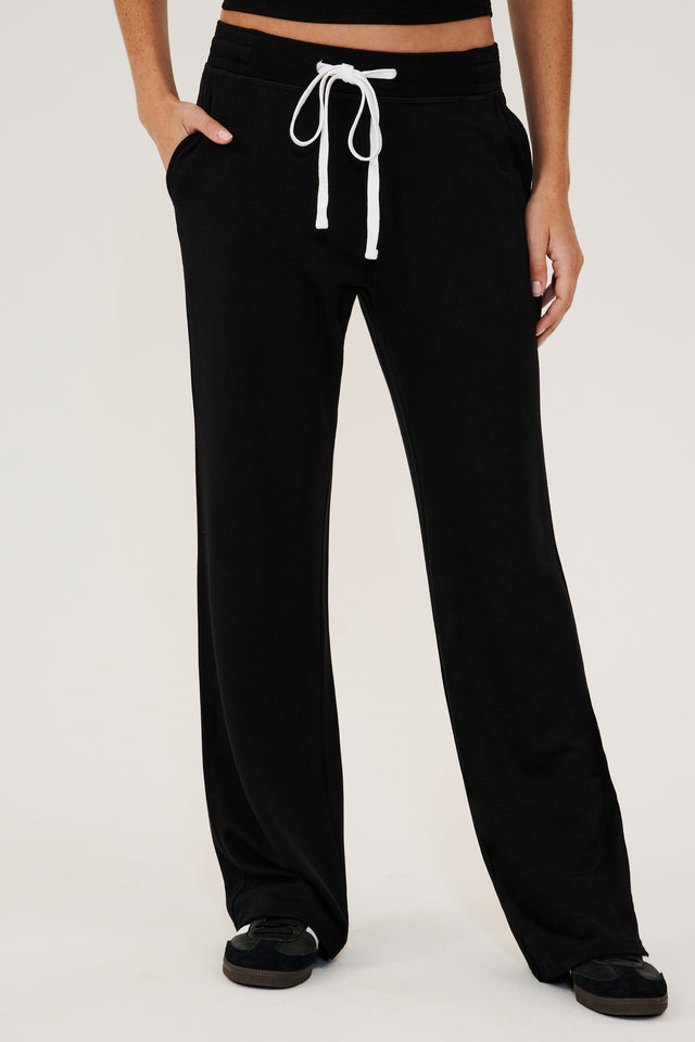 Front view of woman wearing a black high rise wide leg relaxed fit sweatpant with side pockets and white drawstring. Paired with black shoes with white stripes.