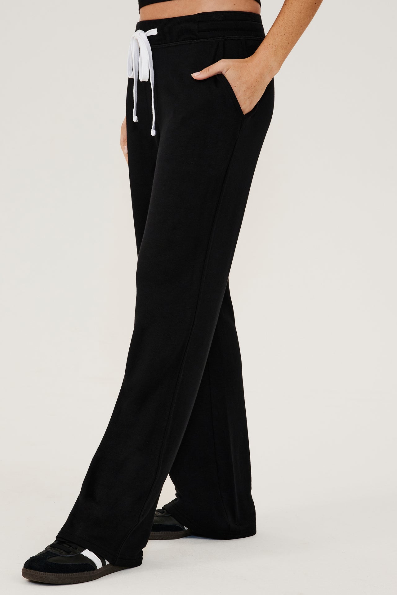 Front side view of woman wearing a black high rise wide leg relaxed fit sweatpant with side pockets and white drawstring. Paired with black shoes with white stripes.