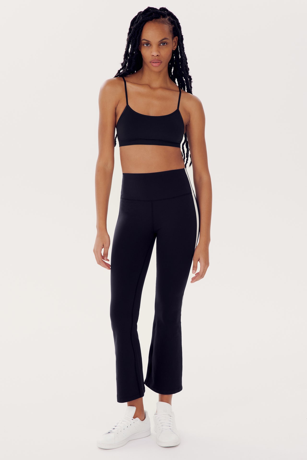A person with long braided hair is wearing a black sports bra made from spandex, SPLITS59 Raquel High Waist Crop - Black/White, and white sneakers, standing against a plain white background.