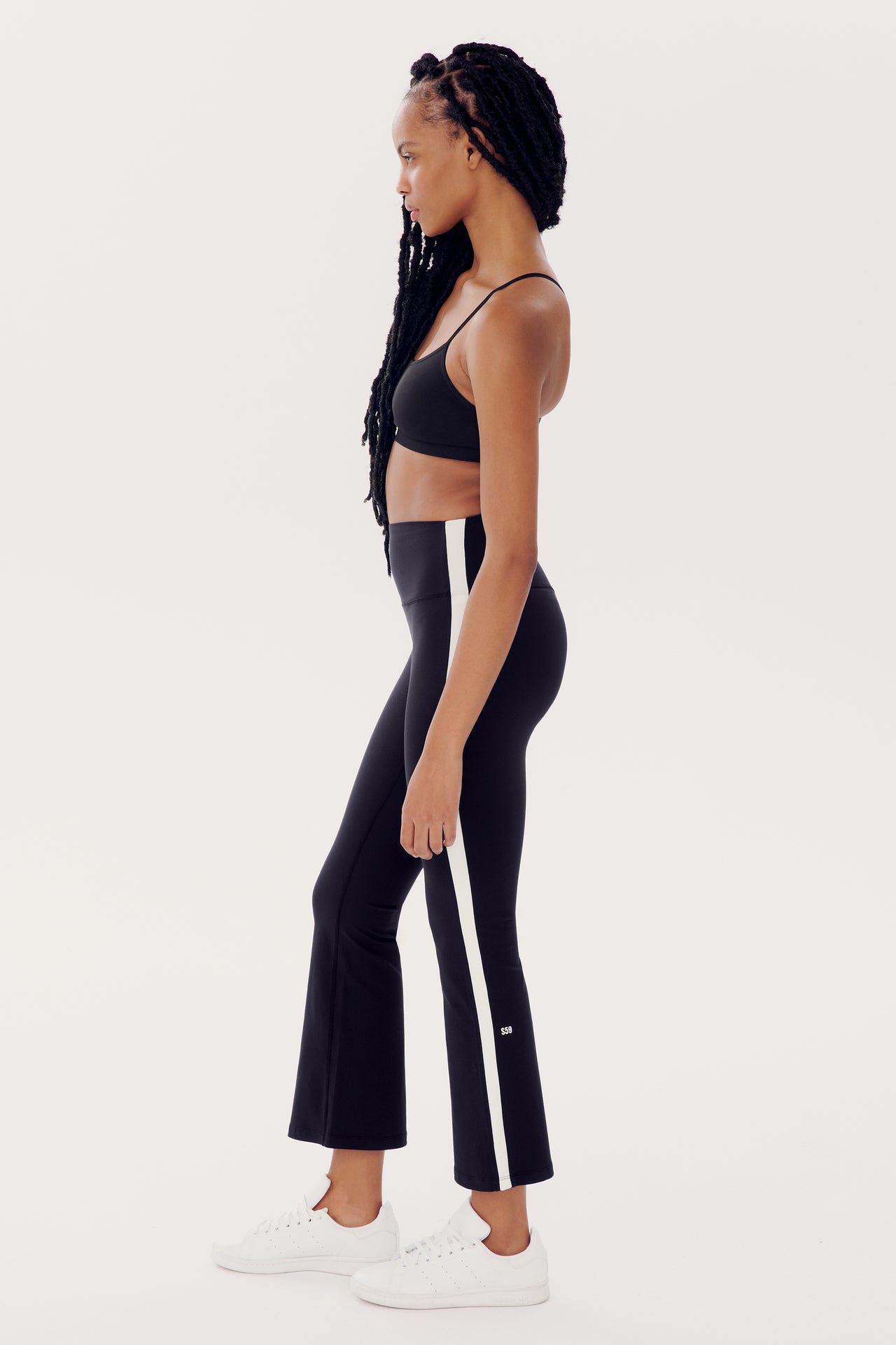 A person wearing a black Raquel High Waist Crop - Black/White by SPLITS59 with a white stripe down the sides, made of stretchy spandex fabric, and white sneakers stands in profile against a plain white background.