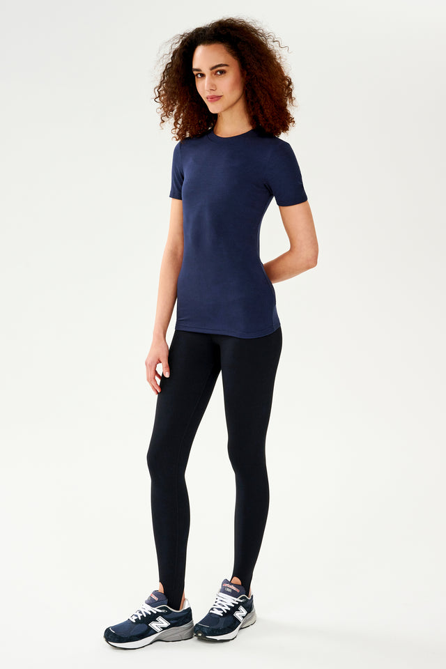 A woman wearing a Louise Rib Short Sleeve in Indigo by SPLITS59 and black leggings, ready for her yoga session.