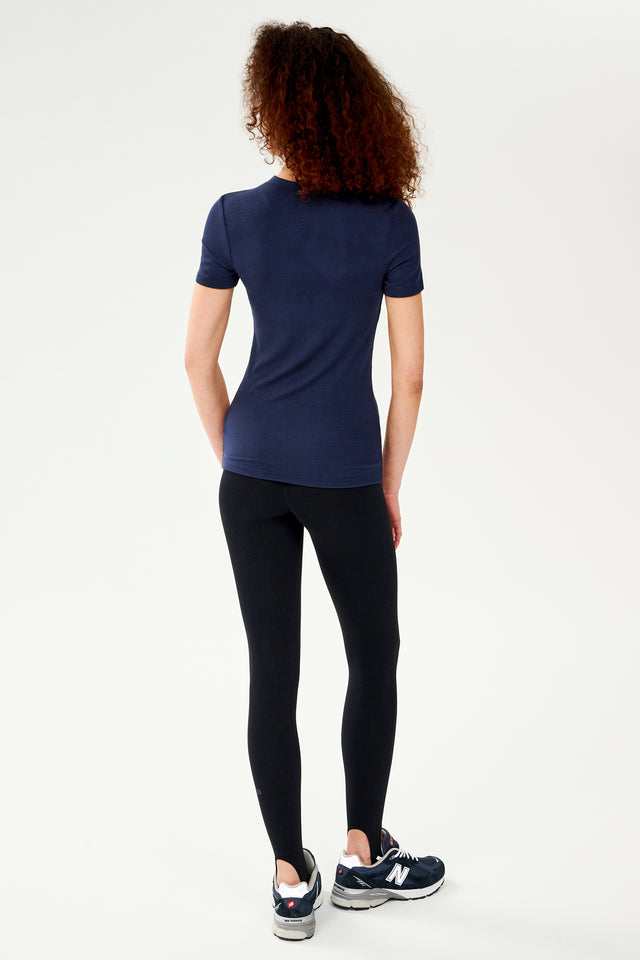 The back view of a woman wearing a SPLITS59 Louise Rib Short Sleeve - Indigo t-shirt and black leggings, ready for her yoga session.