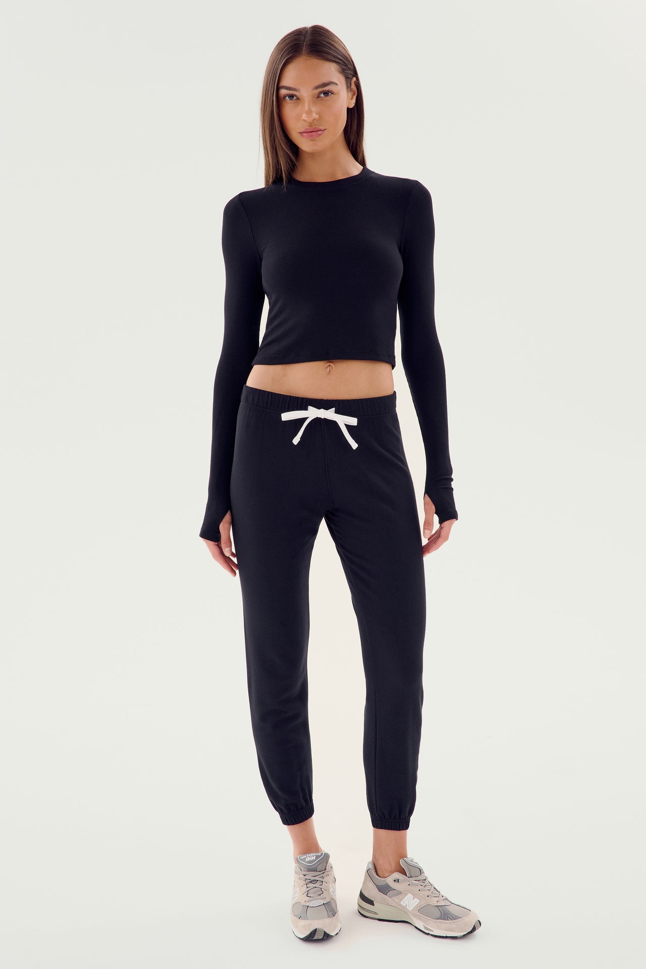 A woman stands facing the camera, wearing a black long-sleeve crop top, Sonja Fleece Sweatpants - Black by SPLITS59 with a white drawstring, and white athletic shoes. She has long, straight hair and a neutral expression, perfect for cool weather workouts.