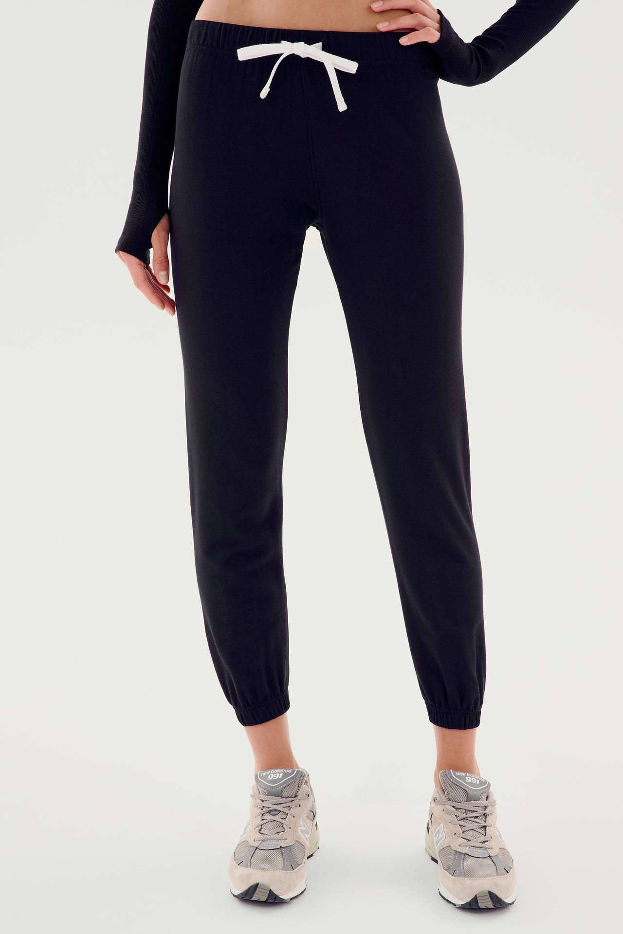 A person is dressed in the Sonja Fleece Sweatpant - Black by SPLITS59, accentuated with a drawstring waist and paired with gray sneakers. These ankle-length sweatpants feature elastic cuffs, making them ideal for cool weather workouts. Their upper body is partially visible, sporting a long-sleeve shirt.