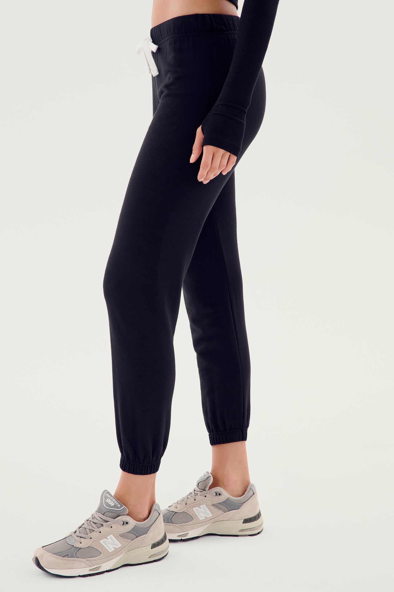 A person stands sideways wearing SPLITS59's Sonja Fleece Sweatpant in black, featuring a white drawstring and paired with grey sneakers, perfect for cool weather workouts, against a plain background.