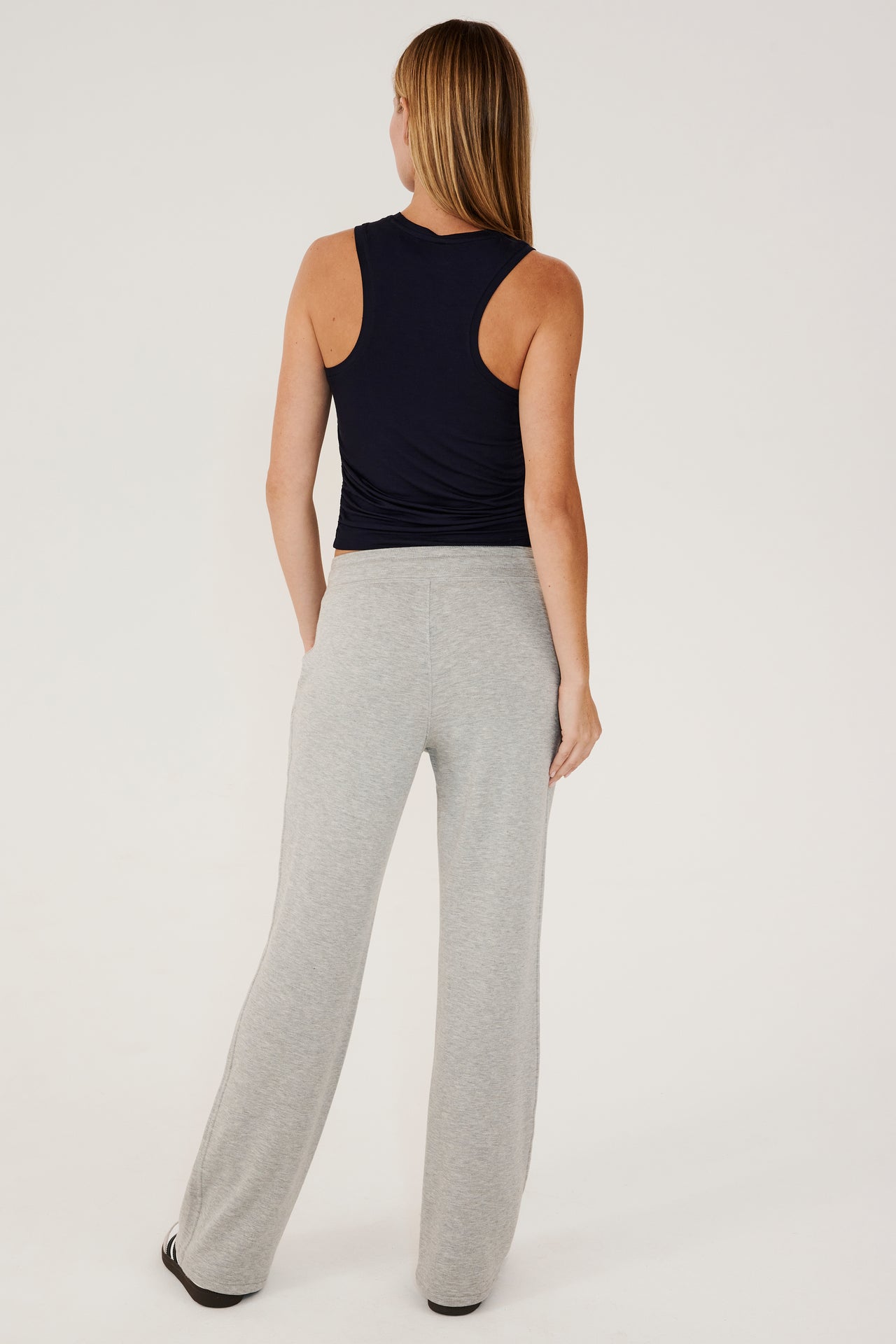 Full back view of woman with straight blonde hair wearing a light gray high rise wide leg relaxed fit sweatpant with side pockets. With black  high neckline racer tank top. Paired with white and grey shoes with black stripes.