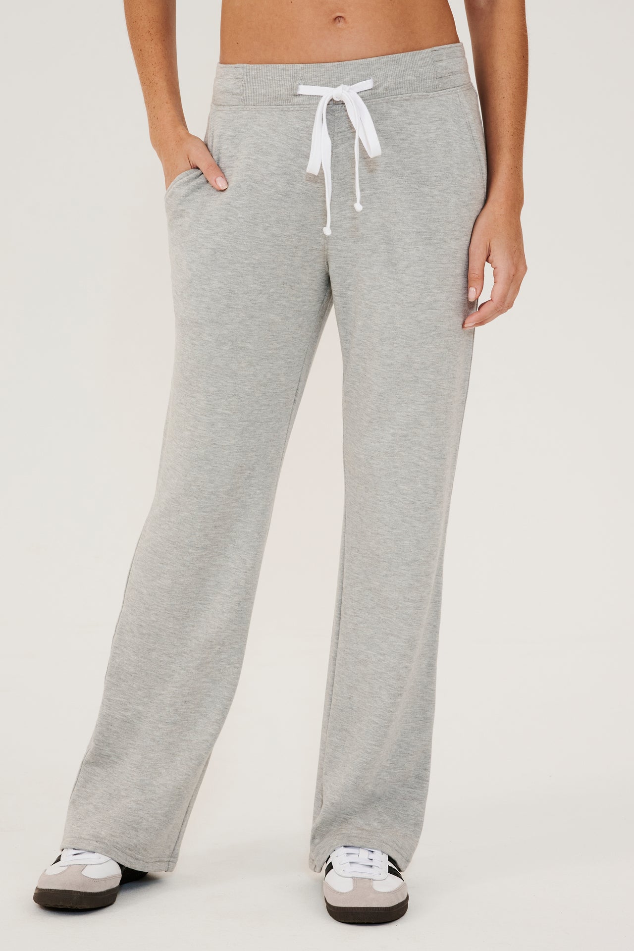 Front view of woman  wearing a light gray high rise wide leg relaxed fit sweatpant with side pockets and white drawstring. Paired with white and grey shoes with black stripes.