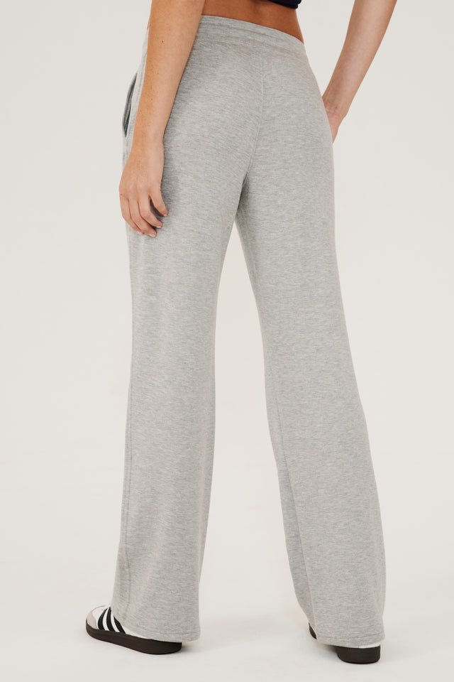 Back side view of woman wearing a light gray high rise wide leg relaxed fit sweatpant with side pockets and white drawstring. Paired with white and grey shoes with black stripes.