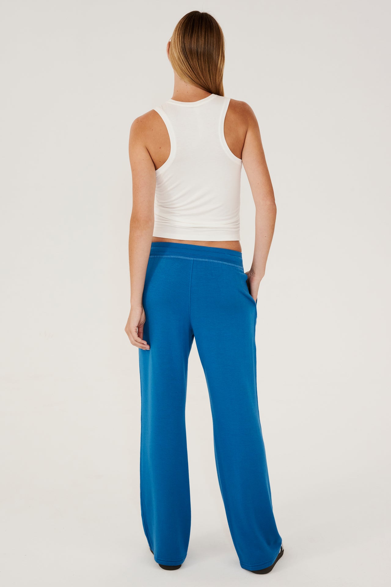 Full back view of woman with straight blonde hair wearing a bright blue high rise wide leg relaxed fit sweatpant with side pockets and white drawstring. With white high neckline racer tank top. Paired with white and grey shoes with black stripes.