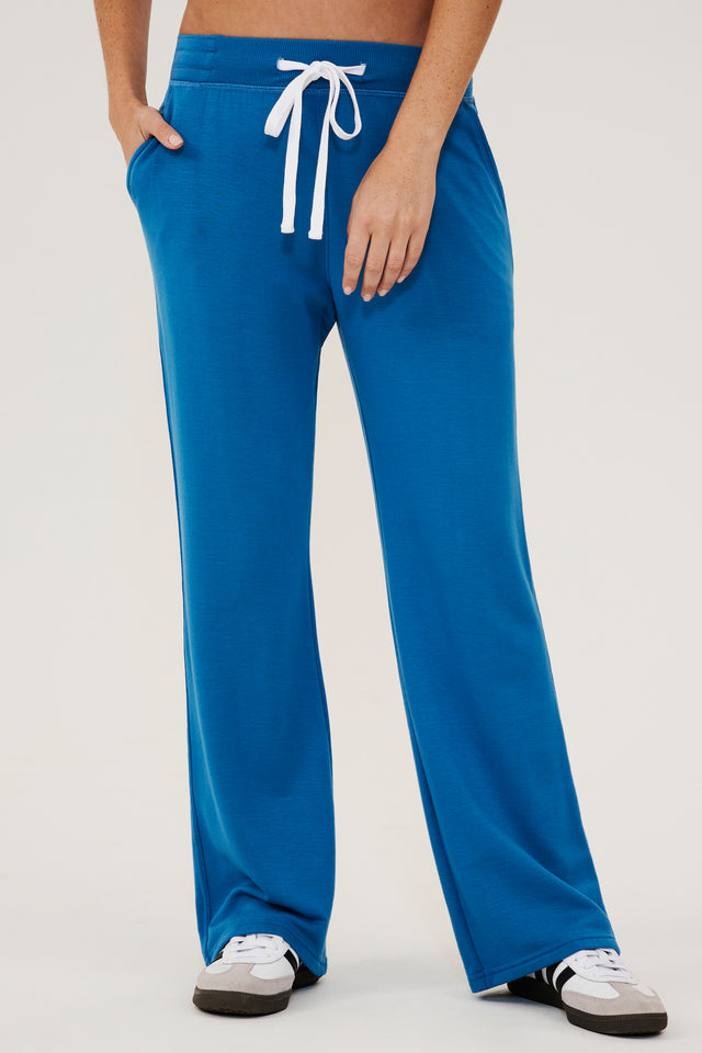 Front view of woman wearing a bright blue high rise wide leg relaxed fit sweatpant with side pockets and white drawstring. Paired with white and grey shoes with black stripes.