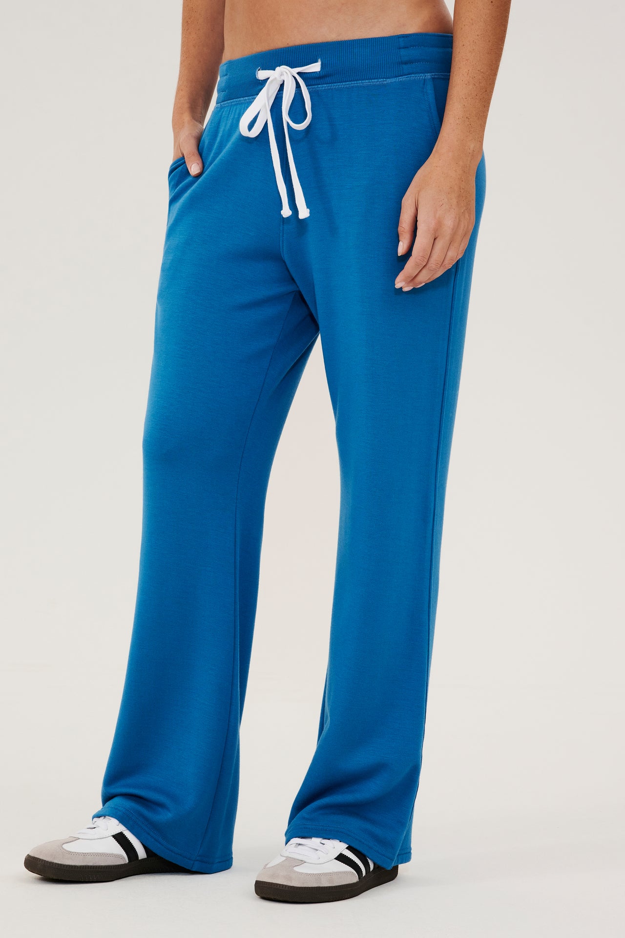 Front side view of woman wearing a bright blue high rise wide leg relaxed fit sweatpant with side pockets and white drawstring. Paired with white and grey shoes with black stripes.