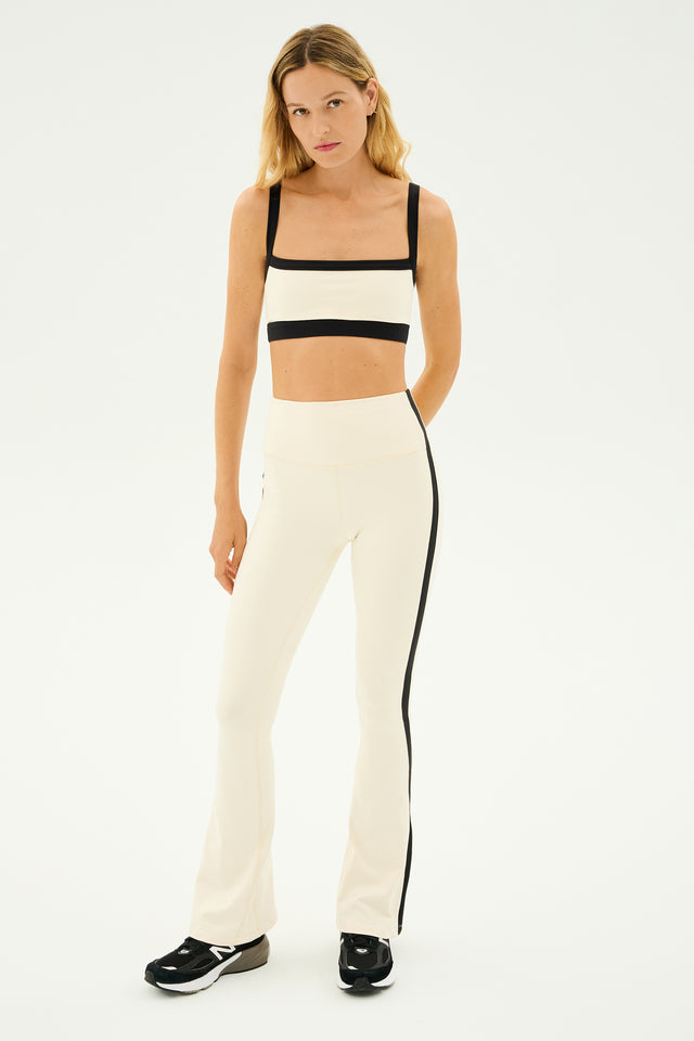 A woman stands against a plain background wearing a two-piece athletic outfit by SPLITS59, featuring the Raquel High Waist Flared Legging in cream with black accents and a matching bandeau top. The ensemble is crafted from 4-way stretch supplex for optimal comfort during any workout.