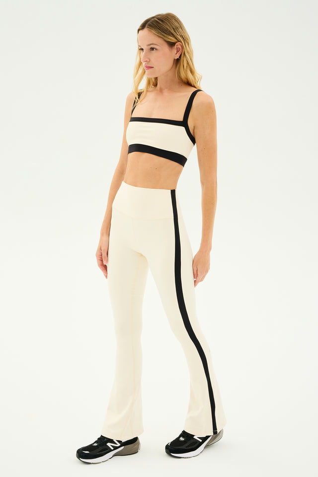 A woman in a beige and black workout outfit stands on a white background, wearing a sports bra and SPLITS59 Raquel High Waist Flared Leggings in Creme/Black. The 4-way stretch supplex material provides comfort, complementing her black New Balance sneakers perfectly.