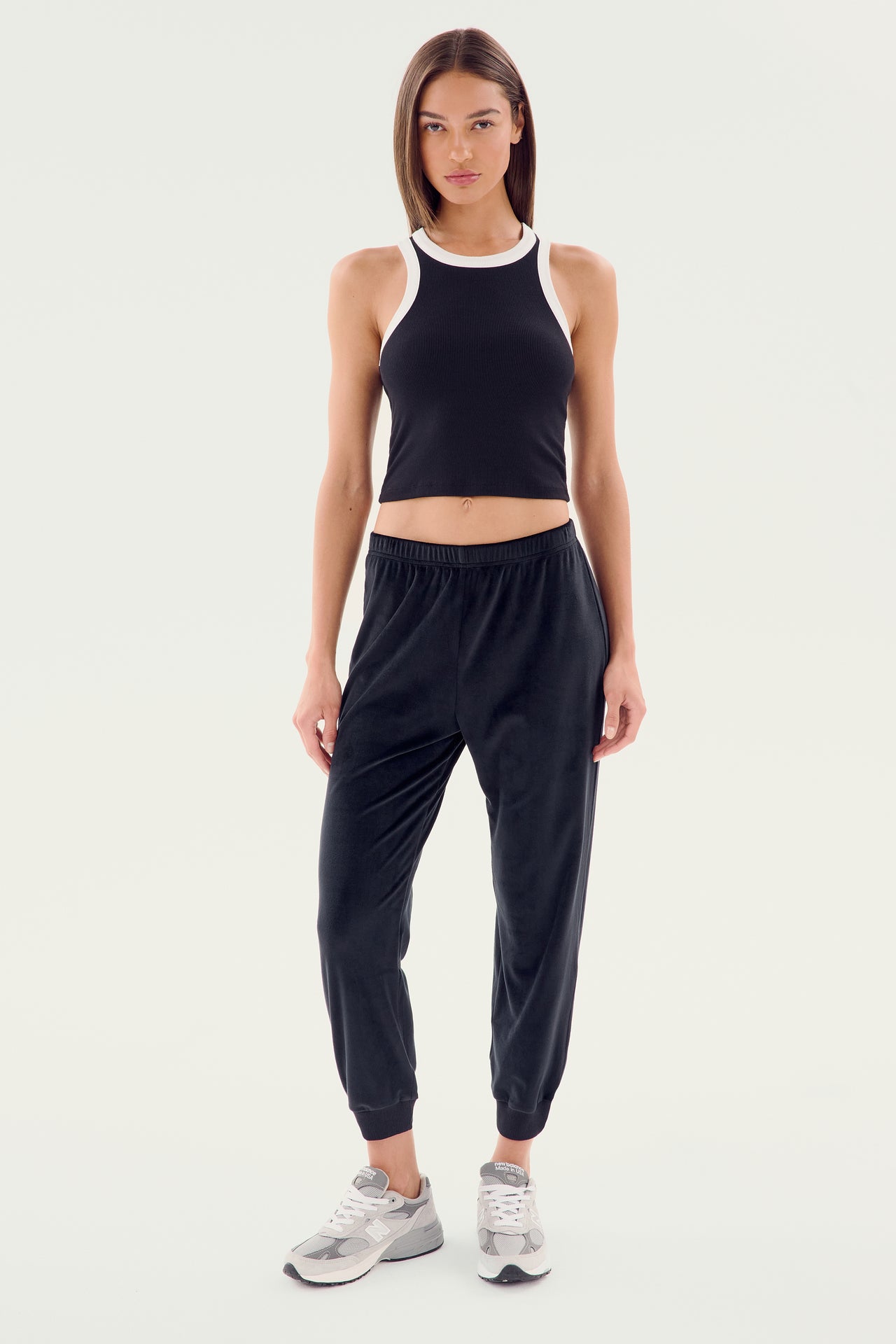 A woman is standing against a plain background wearing the SPLITS59 Kiki Rib Crop Tank - Black/White, black jogger pants, and gray sneakers, perfect for gym workouts.