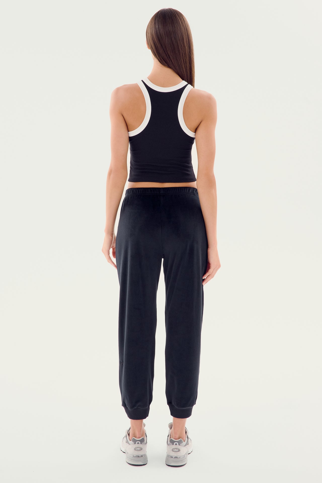 A person stands facing away wearing the SPLITS59 Kiki Rib Crop Tank in black with white trim, black jogger pants, and gray sneakers, ready for an intense gym workout.