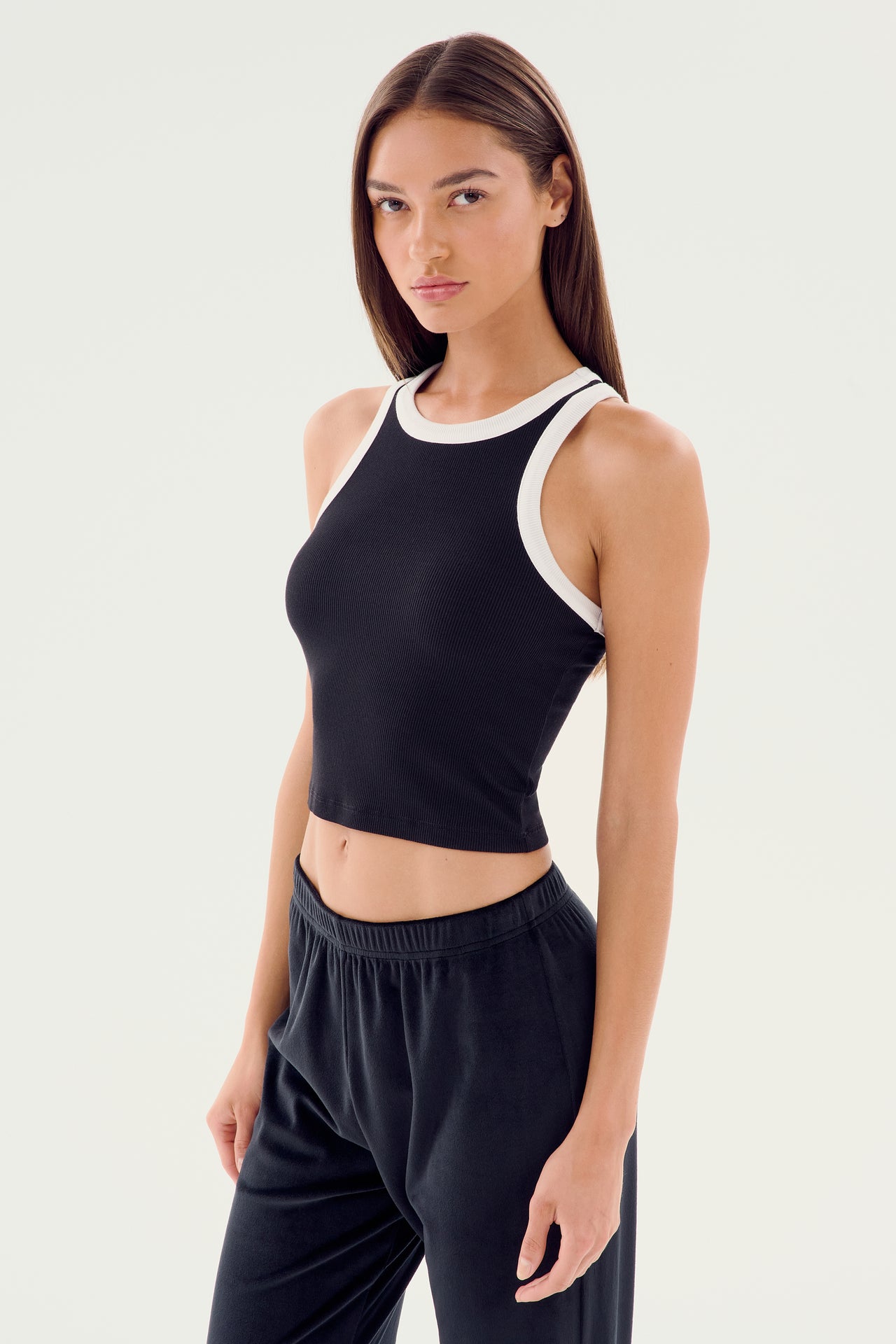 A woman with long, straight hair wears the SPLITS59 Kiki Rib Crop Tank in black and white, paired with black pants, standing against a plain white background.