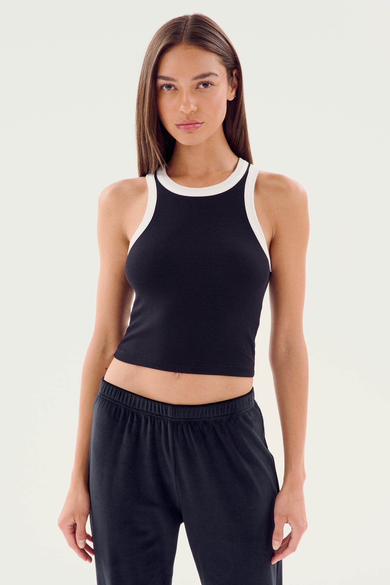 A woman wearing a SPLITS59 Kiki Rib Crop Tank in black and white stands against a plain background, looking directly at the camera. She’s also sporting black pants that are perfect for gym workouts.