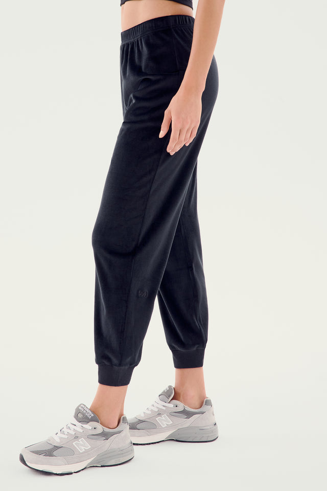 Person wearing SPLITS59's Rhys Velour Jogger in black and gray running shoes, standing against a plain white background. The casual wear outfit emphasizes both comfort and style, with only the lower half of the body visible.