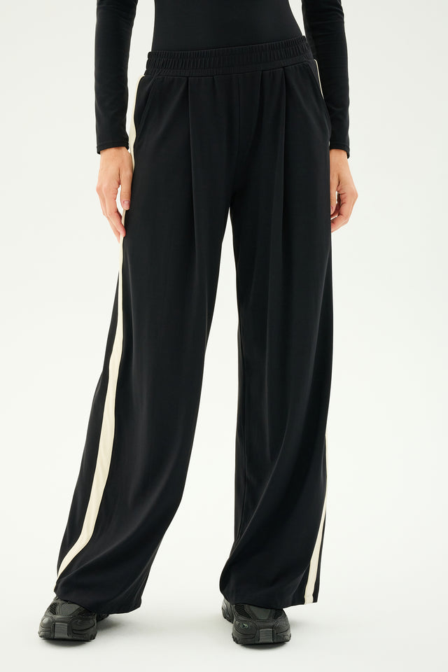 A person dressed in black SPLITS59 Luca Airweight Trousers with Stripe in Black/White, paired with a black top and black shoes, is standing against a plain background. These versatile trousers are known for their style and comfort.