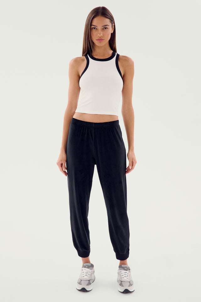 A woman with long hair stands facing forward, wearing the Kiki Rib Crop Tank - White/Black by SPLITS59, black sweatpants, and grey sneakers against a plain background.