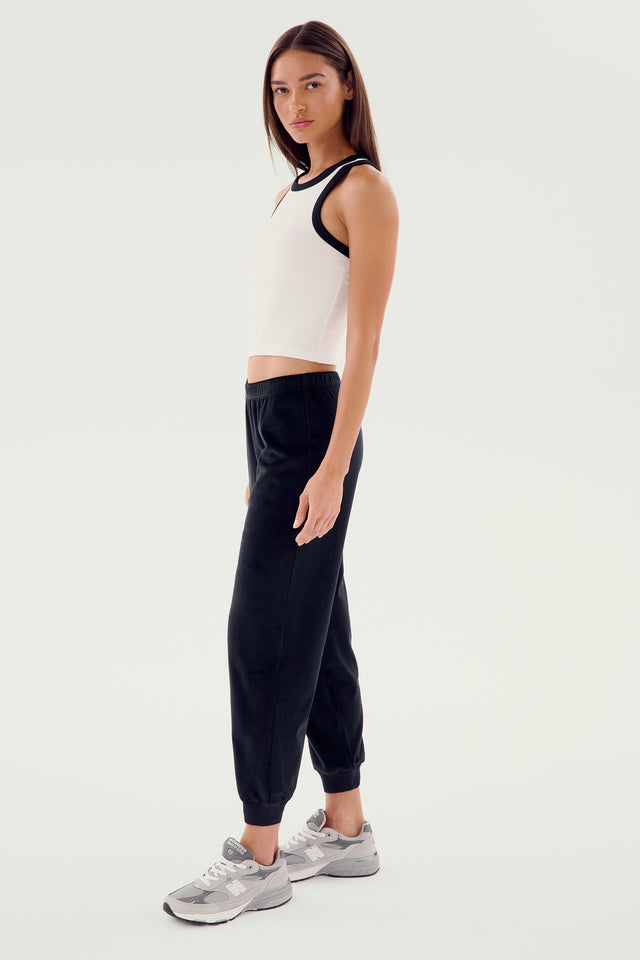 A person with shoulder-length hair wearing the SPLITS59 Kiki Rib Crop Tank in white, paired with black pants and gray sneakers, stands against a plain background, looking ahead.