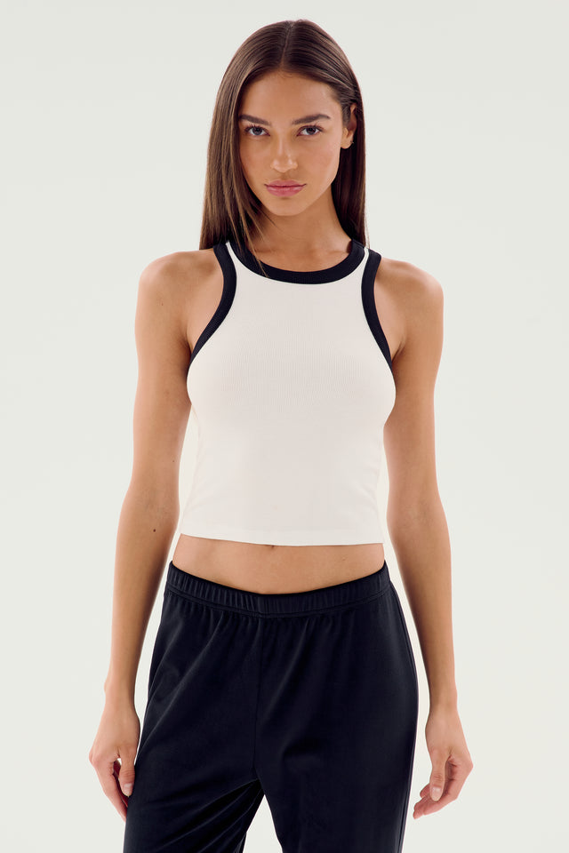 A person with long, straight hair stands facing the camera, wearing the SPLITS59 Kiki Rib Crop Tank in white with black trim and black pants, evoking a gym workout vibe against a plain background.