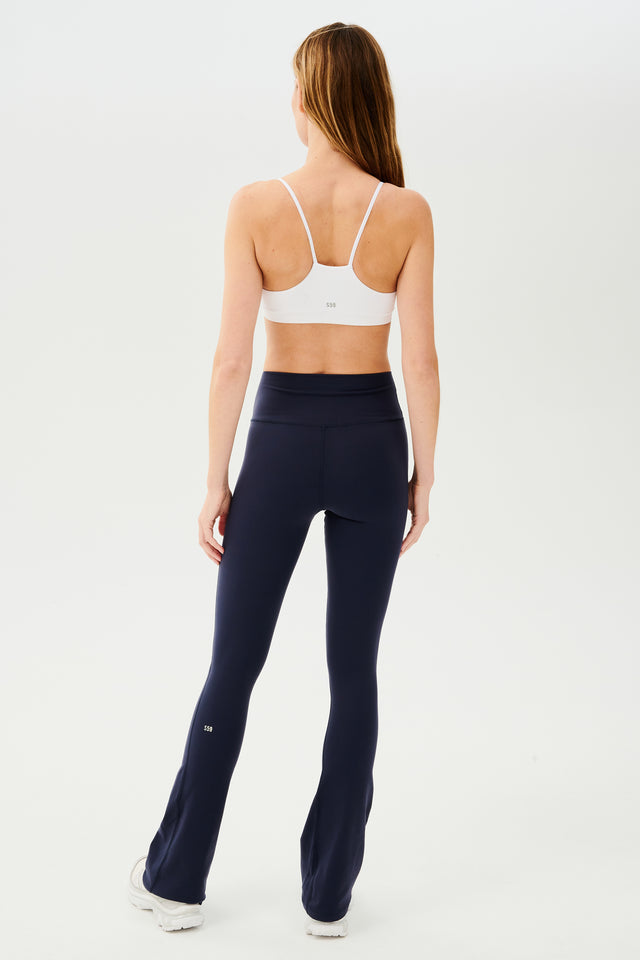 Full back view of woman with straight dark hair wearing dark blue high waist below ankle length legging with wide flared bottoms and white S59 logo on back of left calf. With white racerback bra with spaghetti straps. Paired with white shoes.
