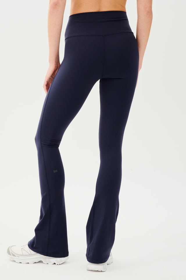 Back view of woman wearing dark blue high waist below ankle length legging with wide flared bottoms and white S59 logo on back of left calf. Paired with white shoes.