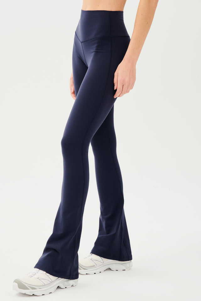 Front side view of woman wearing dark blue high waist below ankle length legging with wide flared bottoms. Paired with white shoes. 