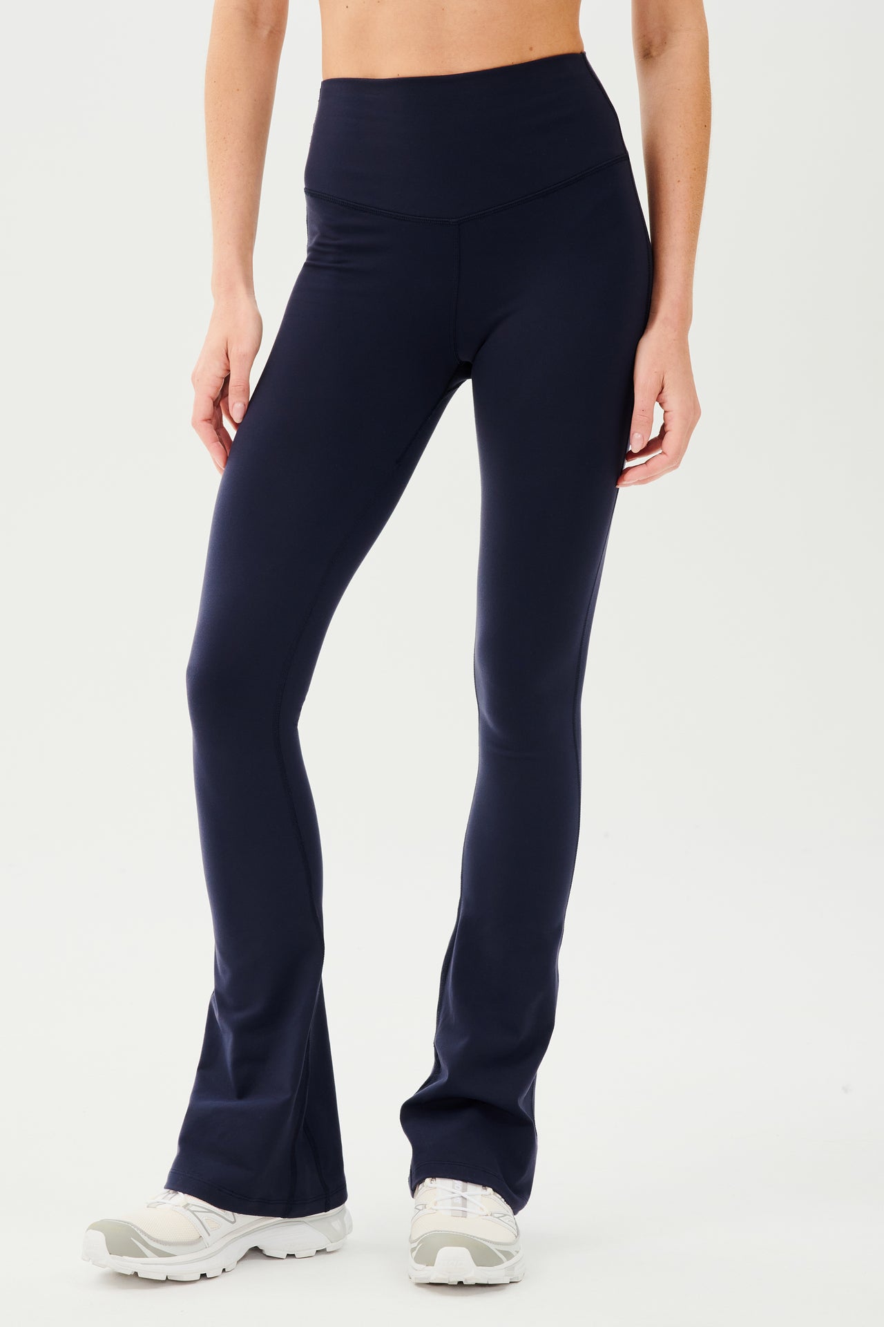 Front view of woman wearing dark blue high waist below ankle length legging with wide flared bottoms.Paired with white shoes. 