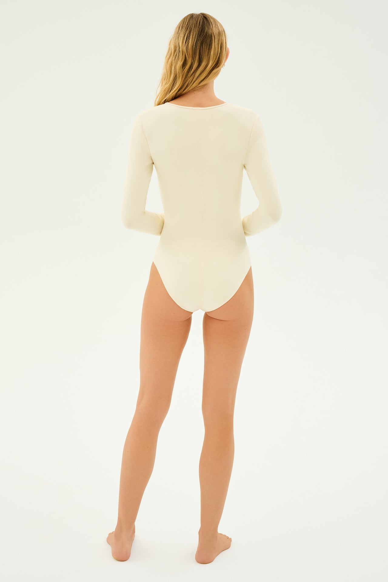 A person with long hair is standing barefoot, facing away, wearing the Airweight Scoop Neck Bodysuit in creme by SPLITS59 against a plain background.