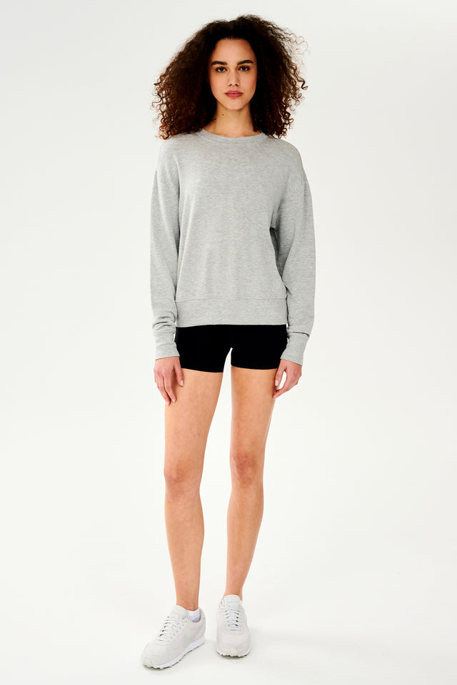 Full front side view of woman with curly dark brown hair wearing a light grey crewneck sweatshirt and black shorts paired with cream shoes