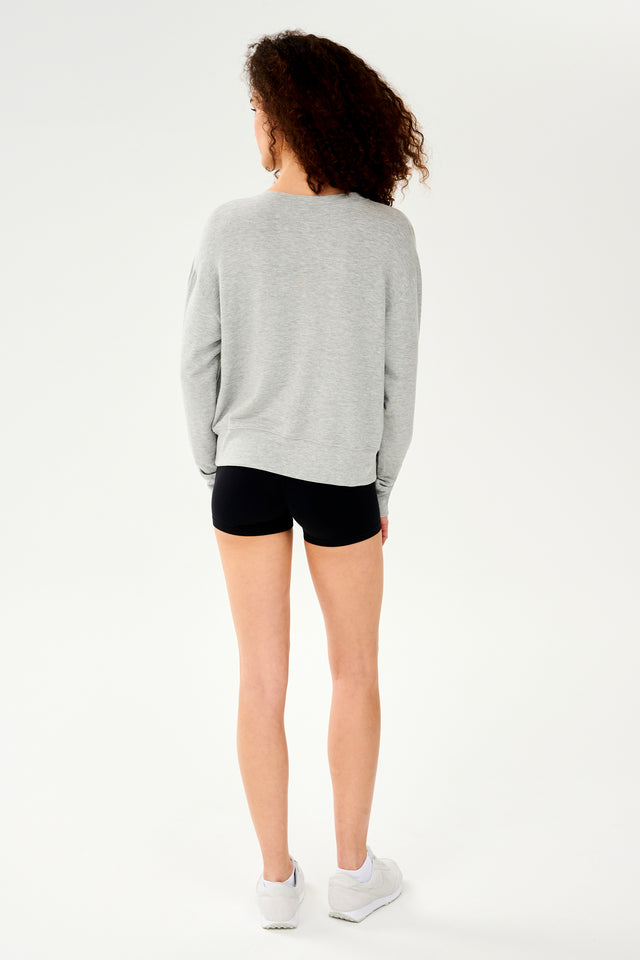 Full back view of woman with curly dark brown hair wearing a light grey crewneck sweatshirt and black shorts paired with cream shoes