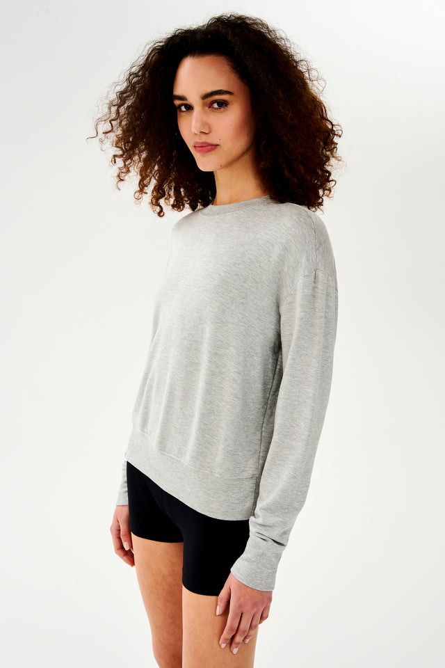 Front side view of woman with curly dark brown hair wearing a light grey crewneck sweatshirt and black shorts 