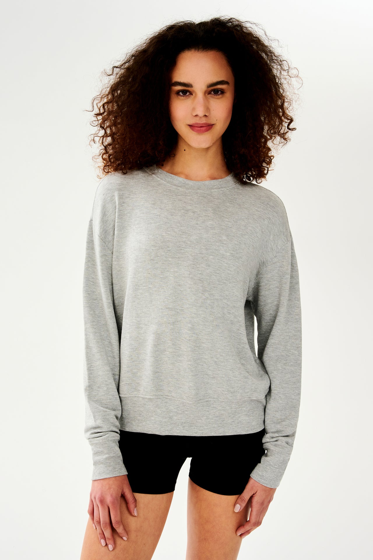 Front side view of woman with curly dark brown hair wearing a light grey crewneck sweatshirt and black shorts 